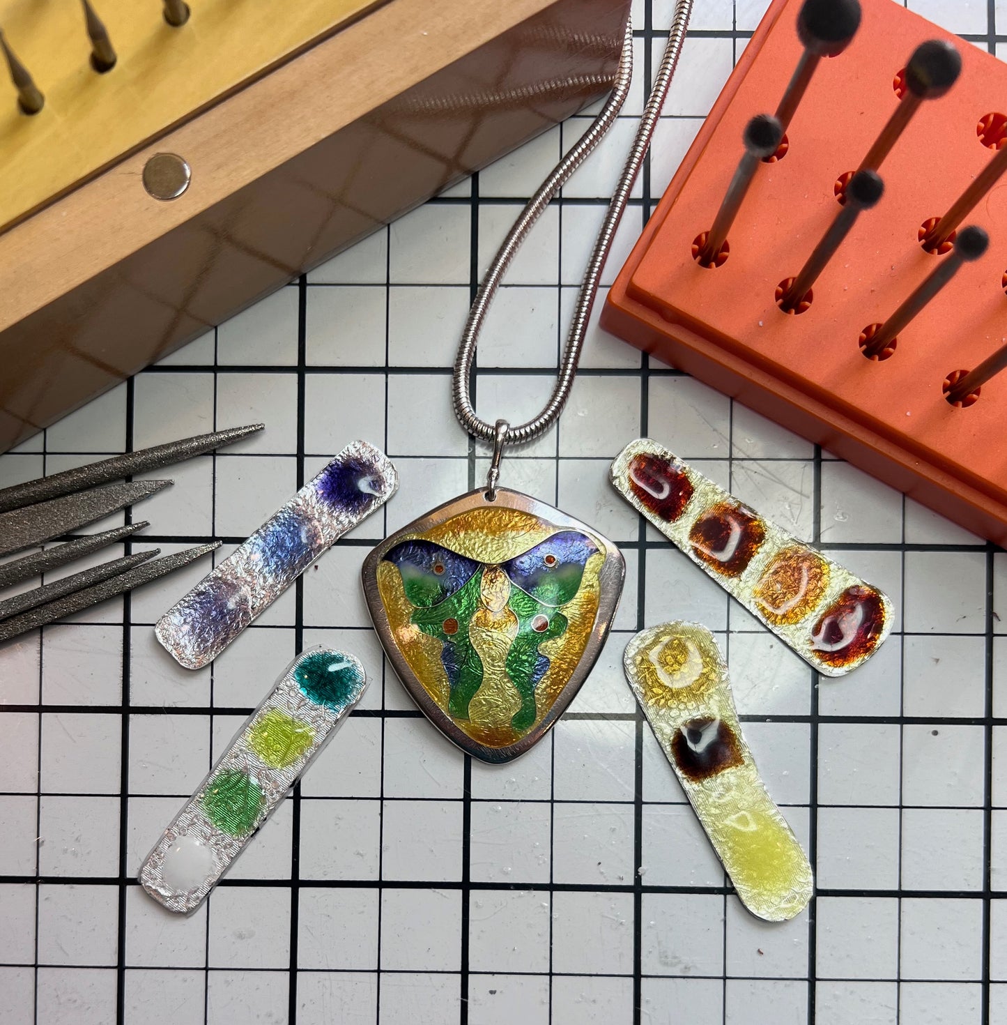 Luna moth enamel pendant on a desk showing enamel colored samples and other jewelry tools