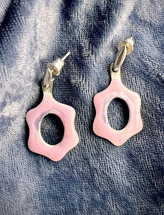 Pair of pink enamel earrings with a cutout center, shaped like a six-petal flower, on a blue textured surface.