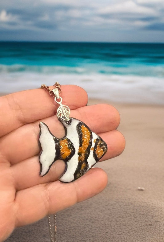 Fish-shaped pendant held in hand with ocean and beach in the background, stripped fish