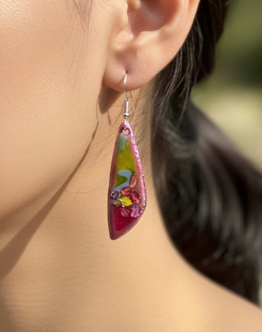 Colorful teardrop-shaped earring worn by a person with a blurred background. Pink and green