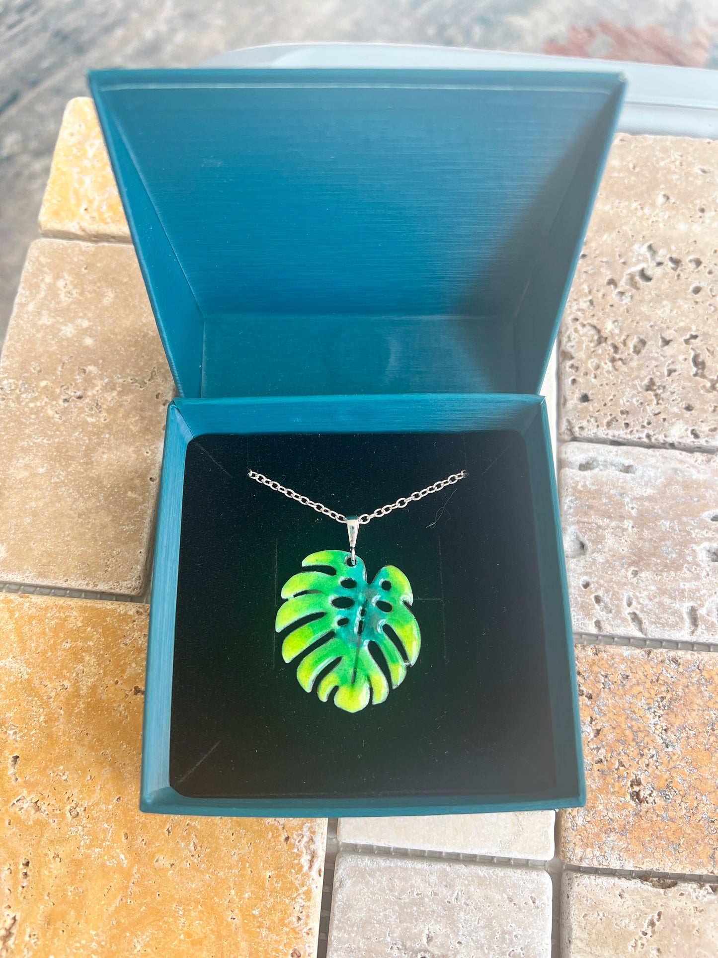 Neon green leaf-shaped pendant in a blue jewelry box on a tiled floor.Monstera leaf