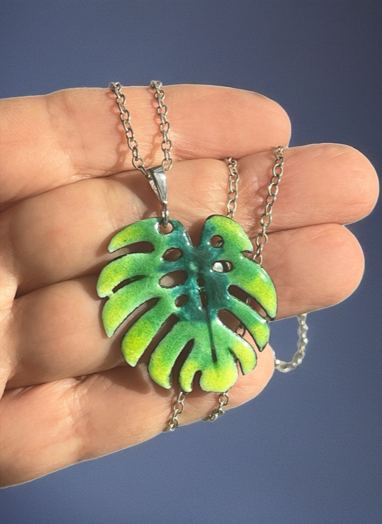Hand holding a necklace with a green leaf pendant against a blue background. Silver Chain