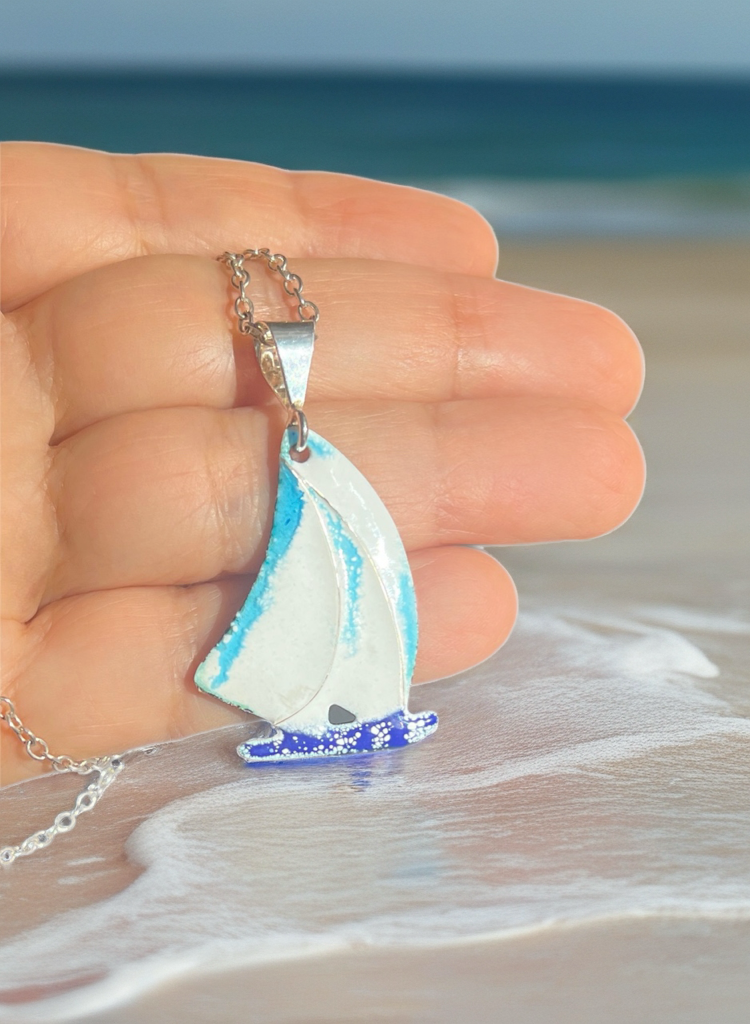 Sailboat pendant on a chain held over a beach with ocean in the background