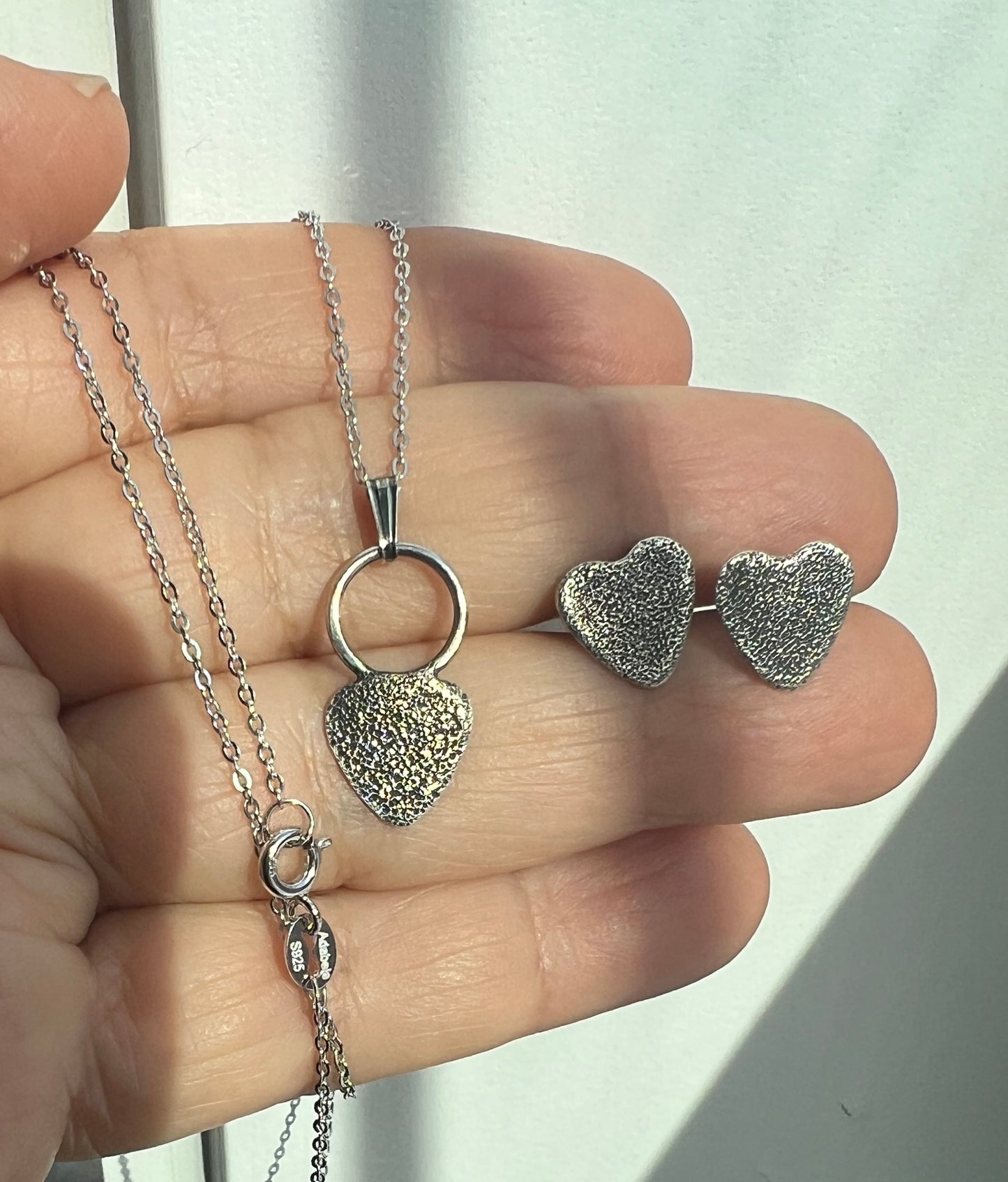 Silver heart-shaped necklace and earrings held in a hand against a light background