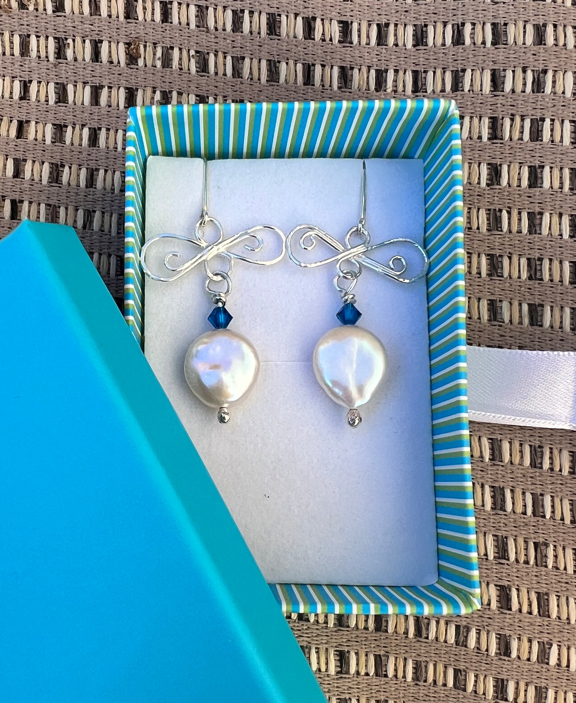 Silver earrings with pearls and blue beads in a striped box on a textured surface.