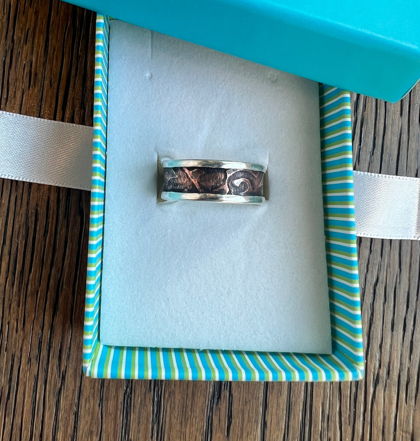 Silver ring in a box with blue and green striped interior on a wooden surface