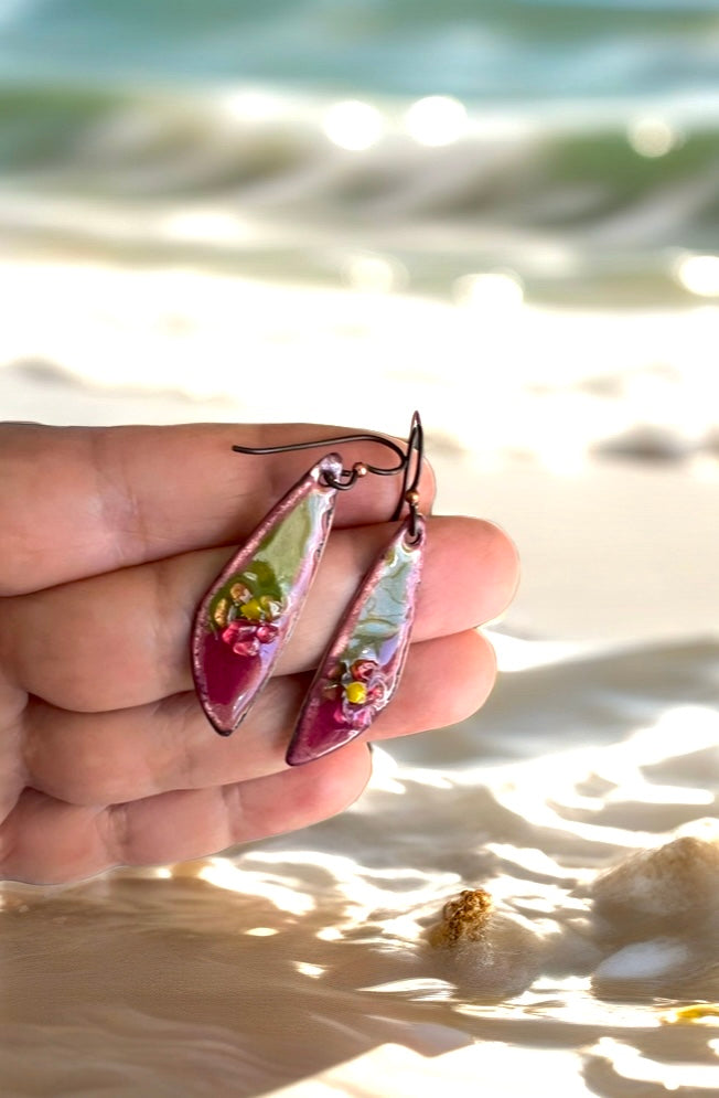Hand holding a pair of floral earrings with a beach background, green and pink earrings