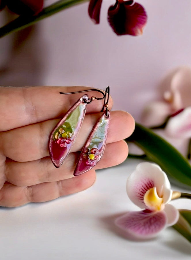 Hand holding a pair of floral earrings with a blurred orchid in the background, pink and green