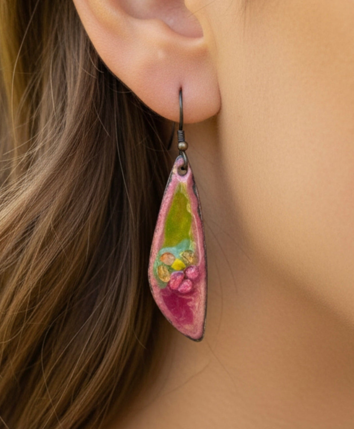 Close-up of an ear wearing a colorful teardrop earring with blurred greenery in the background